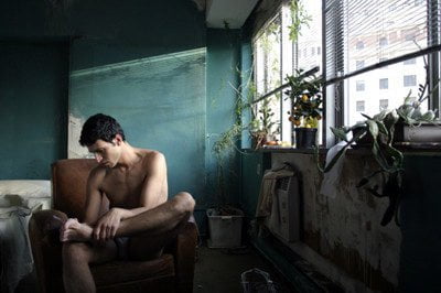 Un joven marroquí, con el torso desnudo, está sentado pensativo en un sillón dentro de una habitación deteriorada. La luz entra por la ventana, iluminando varias plantas y creando un ambiente melancólico.